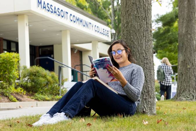 Student reading a textbook outside in front of a tree.